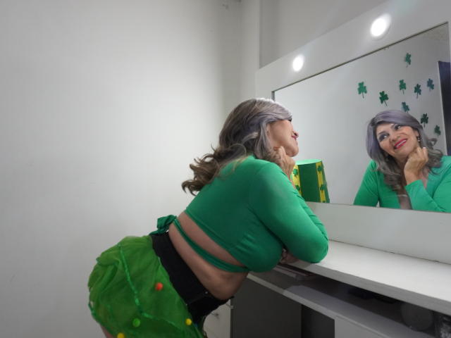 CelesteLincon with green top, green skirt, and grey hair leaning on vanity, looking at mirror, reflecting green eyes, and red lipstick. Boobs slightly visible under green top.