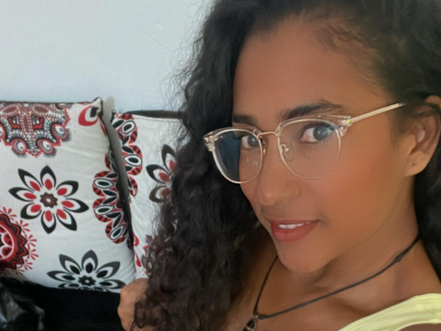 Thannia with curly black hair, tan skin, and glasses, smiling at the camera, showing her small ass and cute nose. She's wearing a yellow top and a black necklace.