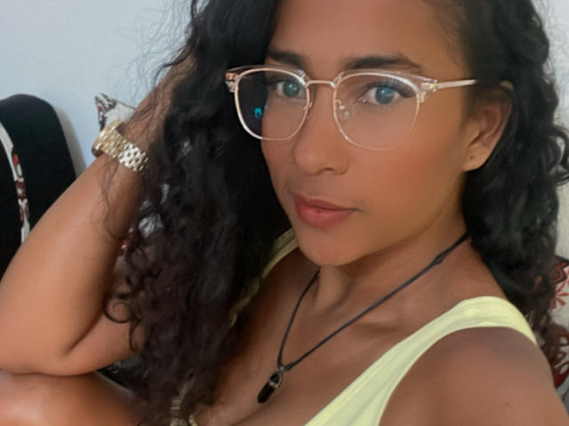Thannia with curly black hair, wearing glasses, a yellow tank top, and a necklace, has tan skin and a serious expression, showing her small tits and a gold wristwatch.