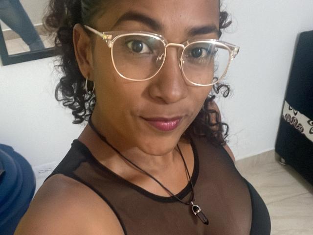 Thannia with curly brown hair, wearing gold glasses, black sheer top, pink lipstick, and a necklace with a small heart pendant, showing her medium brown tits.