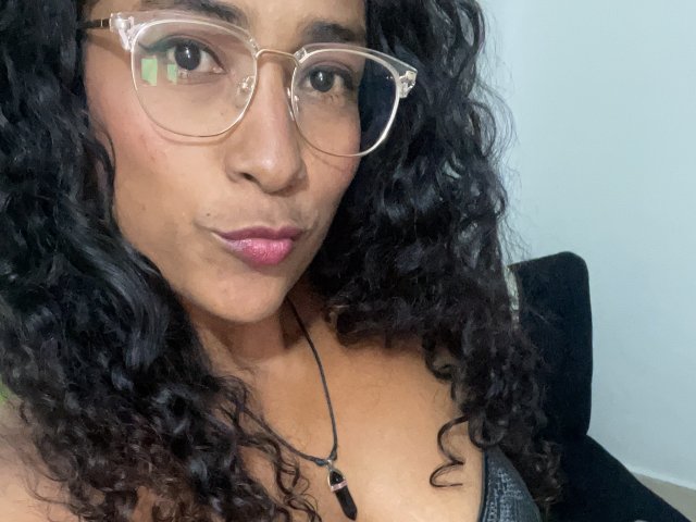 Thannia with curly black hair, pink lipstick, and clear glasses, showing her chest in a black top, small tits, light brown skin, looking at the camera.