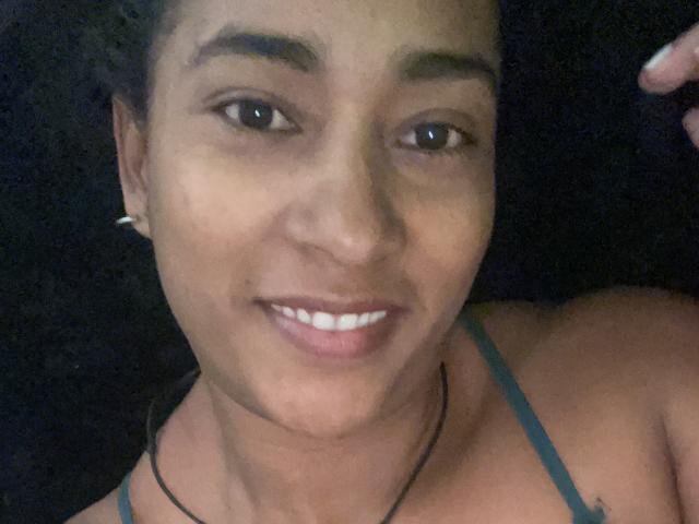 Thannia's face, smiling with white teeth, brown skin, dark hair, small earrings, black necklace, blue strap visible, right hand slightly raised, dark background, casual selfie.
