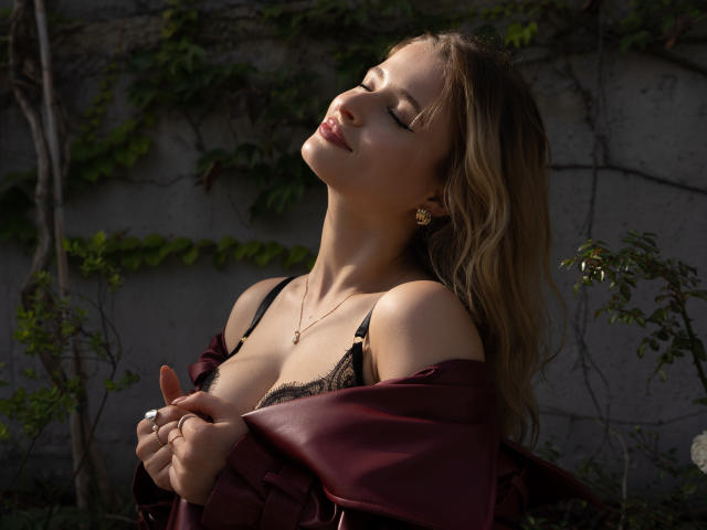 PastelLexy with eyes closed, head tilted back, wearing a black lace bra and maroon jacket, hands holding jacket, gold earrings, sunlit, wavy blonde hair, soft smile.