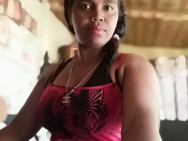 ElenaSarah with dark skin, black braided hair, red and black patterned tank top, black bra, silver necklace with cross, medium tits, slightly blurred background, indoor setting.