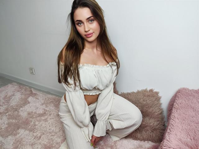 OliviAngel has long brown hair, blue eyes, and is wearing a white off-shoulder crop top with matching pants, kneeling on a pink furry rug. She has small tits and a cute face.