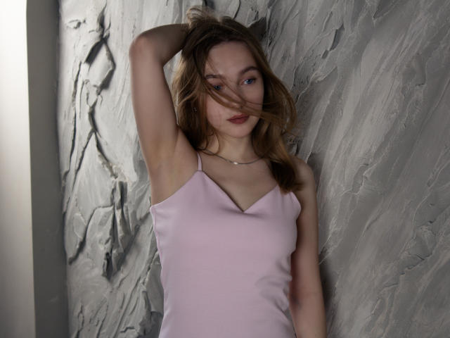 EmiliaMarshall with her arm raised, wearing a pink spaghetti-strap top, showing small tits, against a textured gray wall, with her blonde hair partially covering her face.