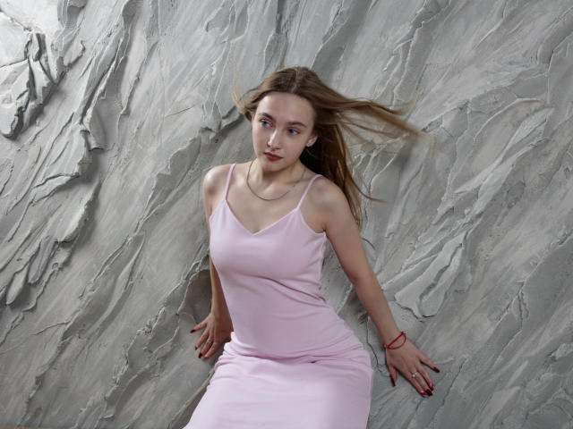 EmiliaMarshall with light brown hair, wearing a pink dress, sitting against a textured gray wall, her arms resting on the surface, looking slightly to the side, wind blowing.