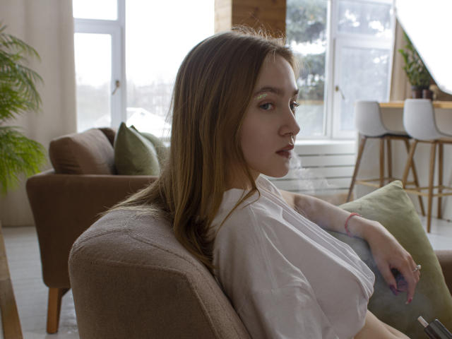 EmiliaMarshall with long brown hair, white shirt, and pink bracelet, sitting on brown sofa, looking to the side, holding phone, green pillows in background, natural light.