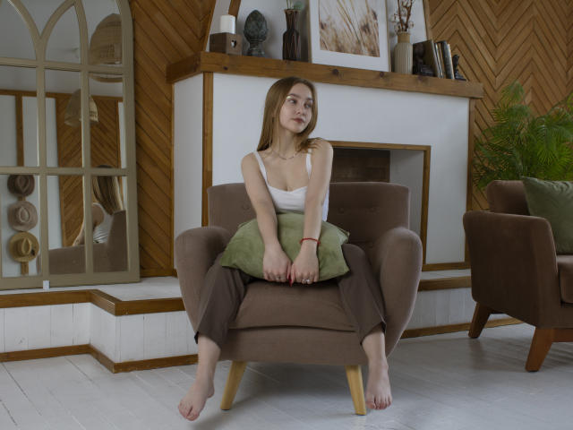 EmiliaMarshall with small tits, sitting barefoot on a brown armchair, wearing a white tank top and brown pants, green pillow on lap, wooden floor, relaxed, smiling.