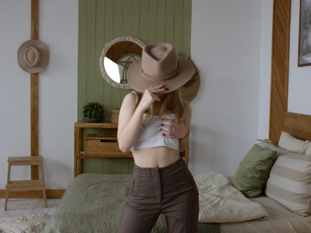 EmiliaMarshall with blonde hair, wearing a brown hat, white crop top, and brown pants, covering her tits with her hand, standing in a bedroom.