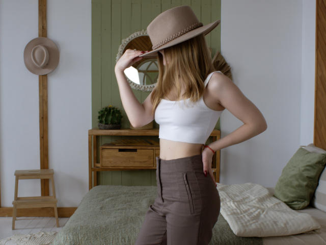 EmiliaMarshall with blonde hair, white crop top, brown pants, and tan hat, touching hat with right hand, showing her small ass and slim body.