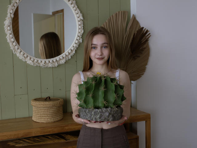 EmiliaMarshall with long brown hair holding a green cactus with spines, wearing a white top and brown skirt, standing in front of a wooden table with a woven basket.