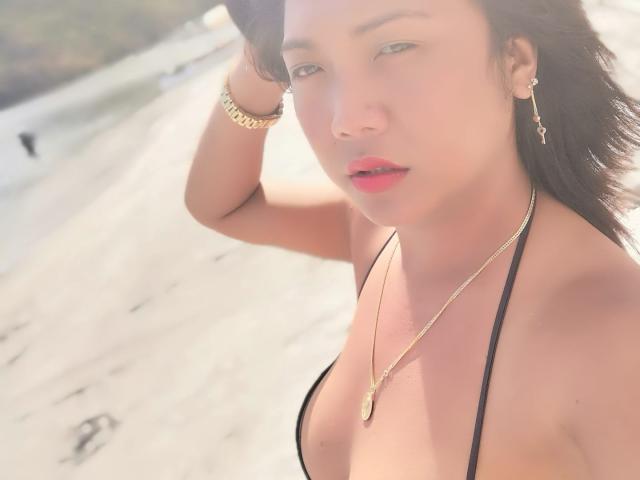 MyObsessionCumTs with light skin, black hair, and pink lips, wearing a black halter top, gold necklace, and bracelet, on a sunlit beach, hand in hair, looking at camera.