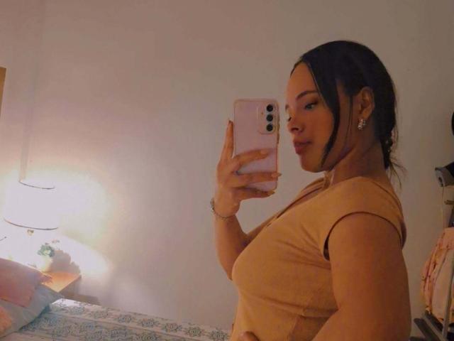JackelineRoset with medium tits, wearing a tight tan top, taking a mirror selfie with a pink phone, dark hair in a ponytail, in a softly lit bedroom.