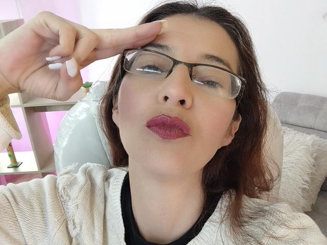 AntonnellaS with glasses, pink lipstick, and white nails, pointing at her forehead, wearing a beige sweater, has medium brown hair, and a slightly pouty expression.