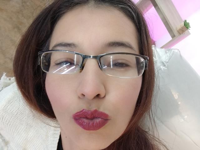 AntonnellaS has red lipstick, pouty lips, and wears glasses with a slight reflection. She has fair skin, brown hair, and is looking at the camera with a subtle smile.