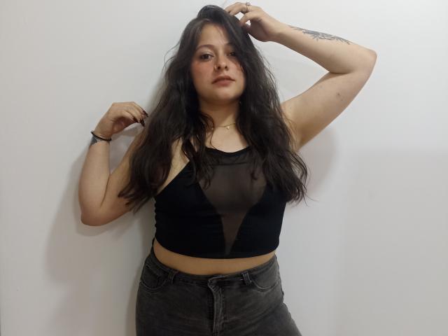AliceWatts with long dark hair, wearing a black sheer top showing her tits, and dark jeans, standing against a white wall, right arm raised, looking at the camera.