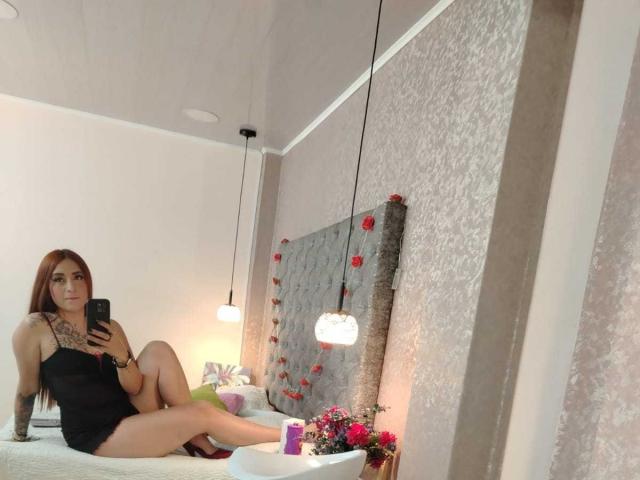 Camile Rouse met lang rood haar, zwarte lingerie, tatoeages op de borst, zittend op een bed, benen gespreid, telefoon in de hand, spiegelbeeld, grijze gestoffeerde hoofdbord met rode rozen, hanglampen..
