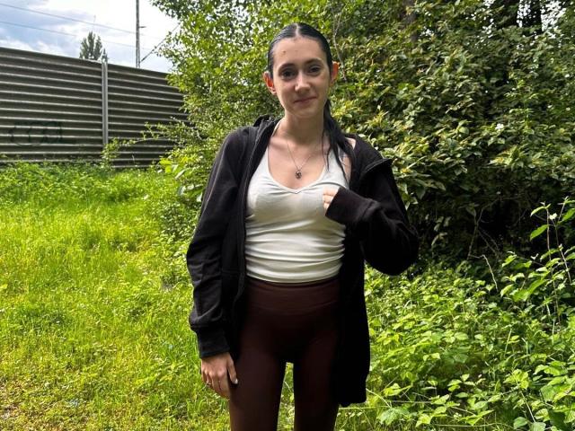 LenaLang-hot with wet dark hair, white tank top, black jacket, and maroon leggings, standing in green grass, touching her chest, looking at camera with slight smile.