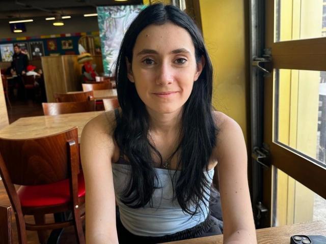LenaLang-hot has black hair, fair skin, small tits, and a subtle smile. She's wearing a white strapless top, sitting at a wooden table in a yellow-walled cafe.