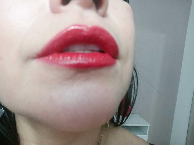 Le labbra rosse e lucide di TamaraFoxy sono leggermente dischiuse, rivelando un accenno dei suoi denti. La sua pelle chiara contrasta con il rossetto rosso acceso. I capelli neri sono parzialmente visibili..