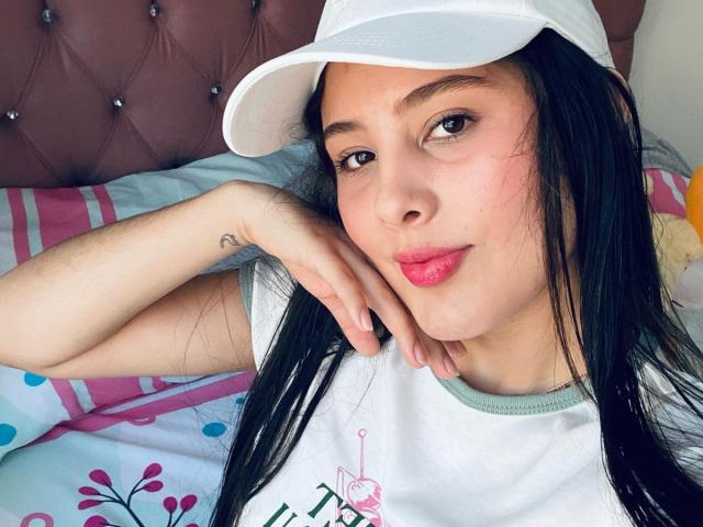 Kathy Stone, met lang zwart haar, die een witte muts draagt en roze lippenstift heeft, ligt op een bed met een tatoeage op haar linkerhand, en ze heeft een schattende, ontspannen glimlach..