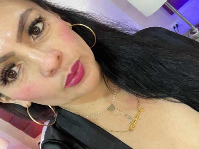 JakyKidman has a cute face with pink lipstick, hoop earrings, and a gold necklace. She has long black hair, brown eyes, and a slight smile. Her tits are partially visible.