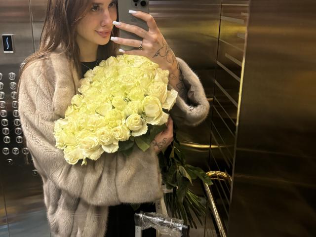 SensualMila with a fluffy gray fur coat, holding a big bouquet of white roses in an elevator, taking a mirror selfie with a subtle smile, tattoos on her right hand.