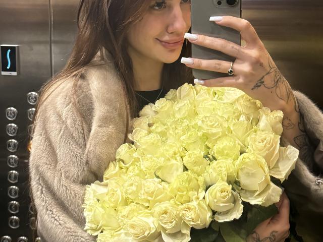 SensualMila mit langen braunen Haaren, weißen Nägeln und einer tätowierten Hand, trägt einen pelzigen Mantel und hält einen Strauß gelber Rosen, während sie ein Selfie in einem Aufzug macht..