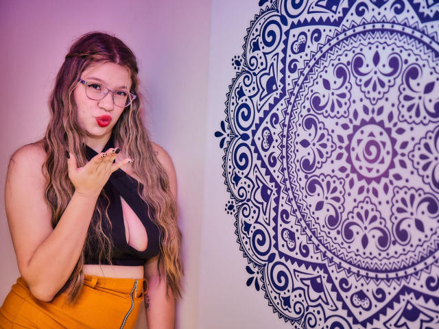 MissGaia with wavy brown hair, glasses, red lipstick, black crop top with cutout, orange high-waisted pants, hand near face, boob outline visible, standing next to intricate blue mandala.