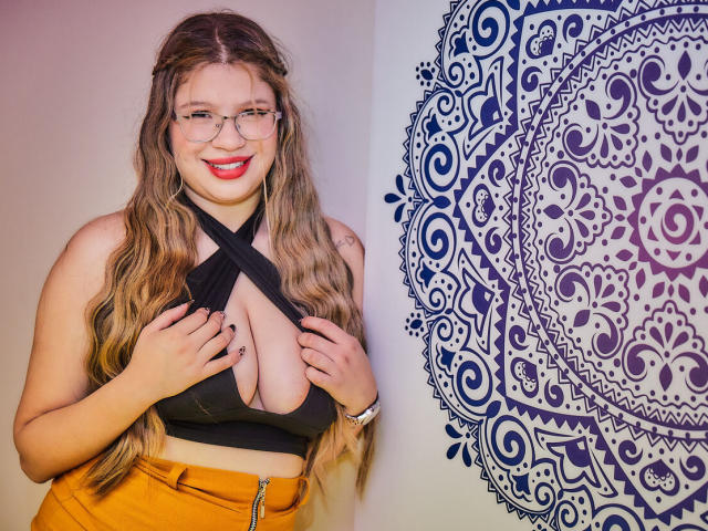 MissGaia with long wavy blonde hair, glasses, red lipstick, black halter top showing her big tits, yellow high-waisted pants, smiling, hands on her chest, mandala wall art.