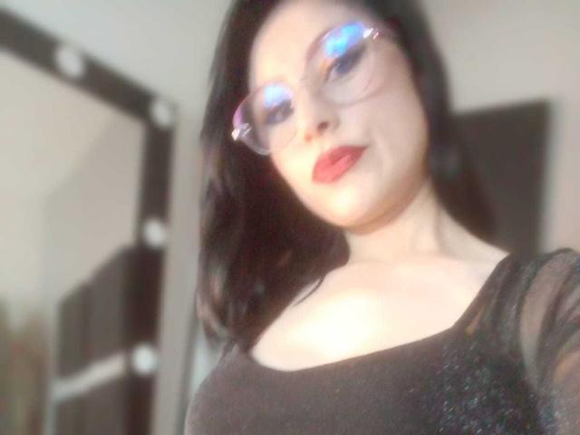 VioletaWhitee with glasses, black hair, red lipstick, and a black sheer top, showing her boobs in a blurry selfie. Her blue-reflecting glasses stand out.