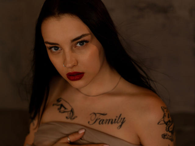 AngelinaSwt with dark hair, red lipstick, tattoos on chest and shoulder, covering her tits with a beige cloth, looking at camera with serious expression.