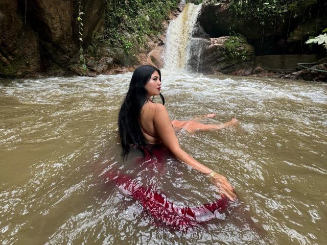 MalakCote, com cabelos longos e pretos, de costas para a câmera, em um rio lamacento, vestindo um vestido vermelho e molhado, sentada, olhando por cima do ombro, com uma cachoeira ao fundo..