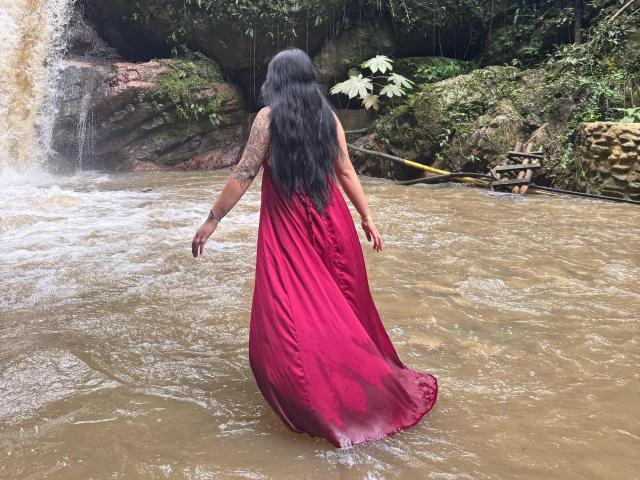 MalakCote stands in muddy water, wearing a long, red, sheer dress that's soaked, with black hair flowing down her back, visible tattoo on her right arm.