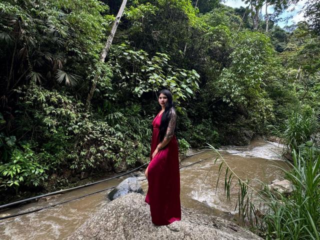 MalakCote stands on a rock in a red dress, tattooed arms, long black hair, and a serious face, with a muddy river and lush green forest behind her.