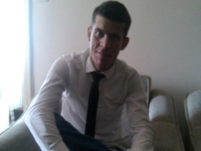 ZionHayes, wearing a white dress shirt and black tie, sits with a slight smile, his dark pants resting on his knees, short brown hair, and light skin.