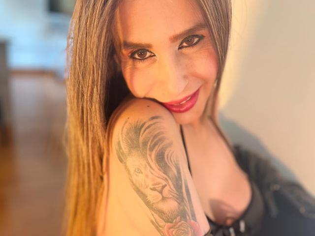 VictoriaJhonnes with long blonde hair, red lipstick, smiling at camera, black top, showing tattooed shoulder of a lion's face, soft lighting, warm brown wood floor, slightly blurred background.