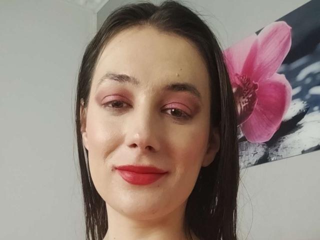 JuliaHuntis has pale skin, brown eyes, pink eyeshadow, red lipstick, straight black hair, and a slight smile. Her ass is not visible. Pink flower painting in background.