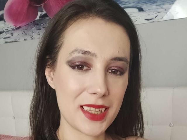 JuliaHuntis has pale skin, dark brown hair, red lipstick, and smoky purple eye makeup, smiling with her teeth visible. Her tits are partially visible in the frame.