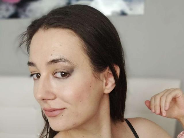 JuliaHuntis has dark brown hair, light skin, small moles on her face, subtle makeup, and a slight smile. Her right hand is raised near her head.