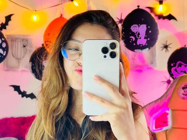 MillyKenner, portant des lunettes, tient un iPhone blanc et prend un selfie. Elle a de longs cheveux bruns, des lèvres roses, porte un haut noir et se trouve dans une pièce décorée sur le thème d'Halloween, avec des décorations orange et noir..