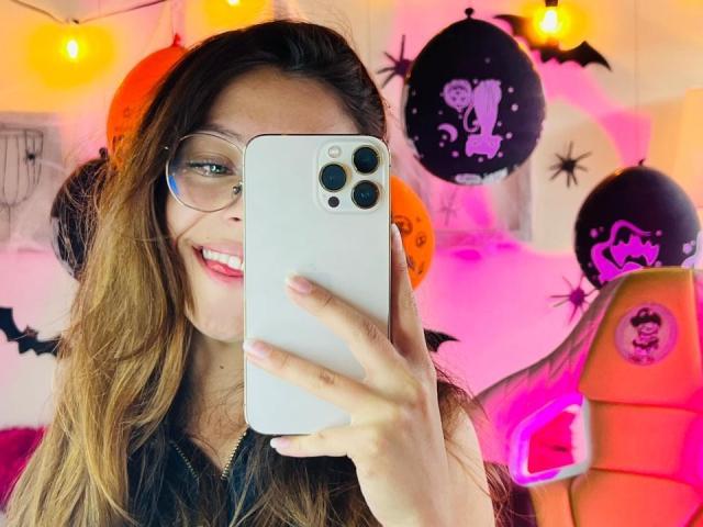 MillyKenner with glasses, smiling, holding a white iPhone, taking a selfie, showing her pink lipstick and brown wavy hair, in a Halloween-themed room with black and orange balloons.