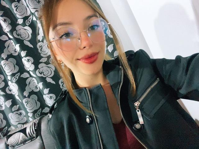 MillyKenner, usando óculos, com um casaco preto, batom vermelho e cabelo loiro, sorrindo para a câmera, mostrando seu rosto fofo e nariz pequeno. Seus seios estão cobertos pelo casaco..