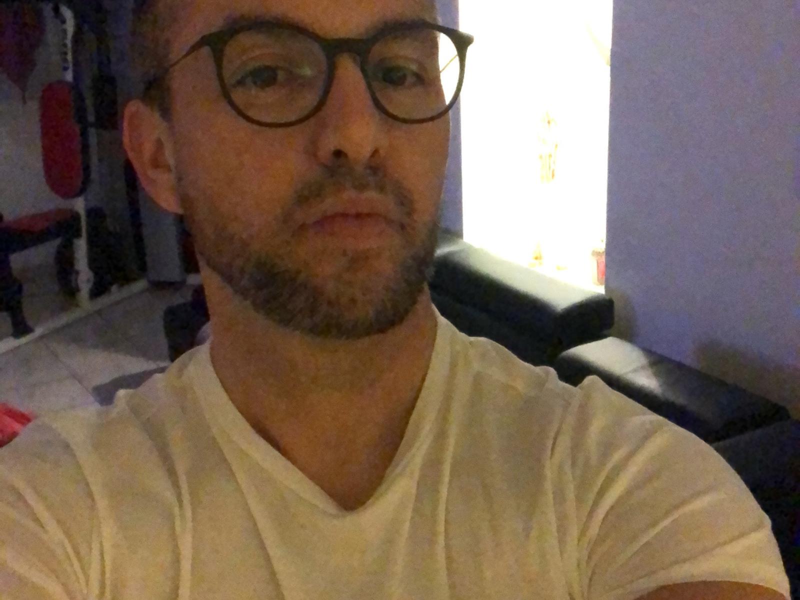 MarvinStar, con gli occhiali, una maglietta bianca, la barba e i capelli corti, guarda verso la telecamera. I suoi glutei sono leggermente visibili sullo sfondo, su un divano in pelle nera..