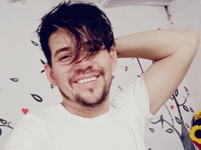 RoxwellTen, con i capelli neri e disordinati, sorride, indossa una camicia bianca, ha un braccio alzato, una piccola barba, orecchini e un viso carino, leggermente buffo. Lo sfondo è decorato con foglie nere e cuori rossi..