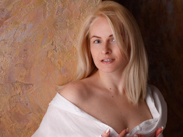AliceeGracee with blonde hair, fair skin, and blue eyes, wearing a white off-shoulder shirt, exposing her small tits, smiling gently, against a textured brown wall.