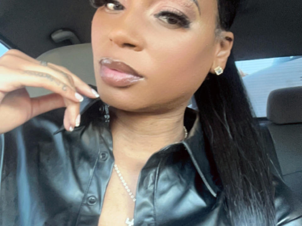 SoReadyForU with dark brown skin, shiny black leather shirt, long black hair, glossy pink lips, subtle eye makeup, small gold earrings, and a delicate necklace, posing in a car.