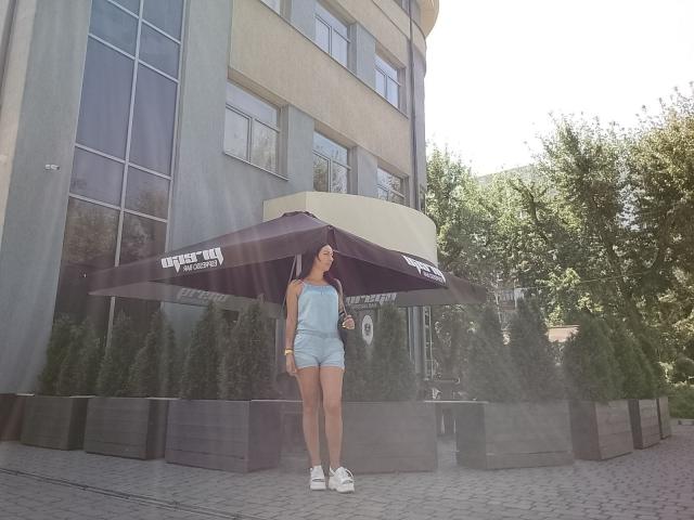 MirandaHott69 staat in de zon, met een grote zwarte paraplu in haar hand, gekleed in een lichtblauwe jumpsuit zonder bandjes en witte sneakers, met een schattige, slanke achterkant en kleine, stevige borsten..