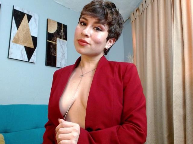 SaraKleine with short brown hair, red blazer, and deep cleavage, showing her bare tits, standing near beige curtains, smiling slightly.
